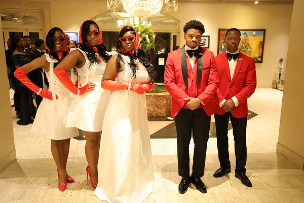 A group dressed elegantly for a formal event, three women in white and two men in red tuxedos.