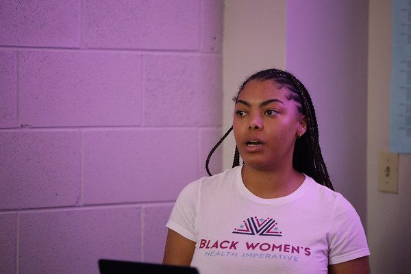 A woman speaking at an event with a purple-lit background.